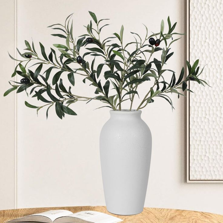 a white vase filled with green leaves on top of a table next to an open book