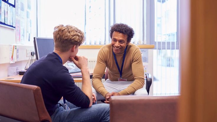 two young men sitting at a table talking to each other in an office setting with laptops on the desk