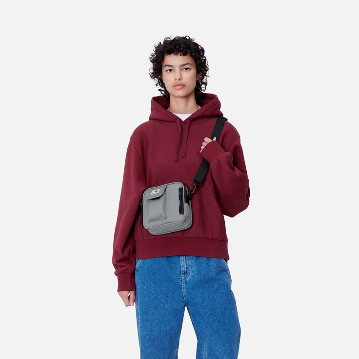 a young man wearing a red sweatshirt and blue jeans, holding a gray fanny bag