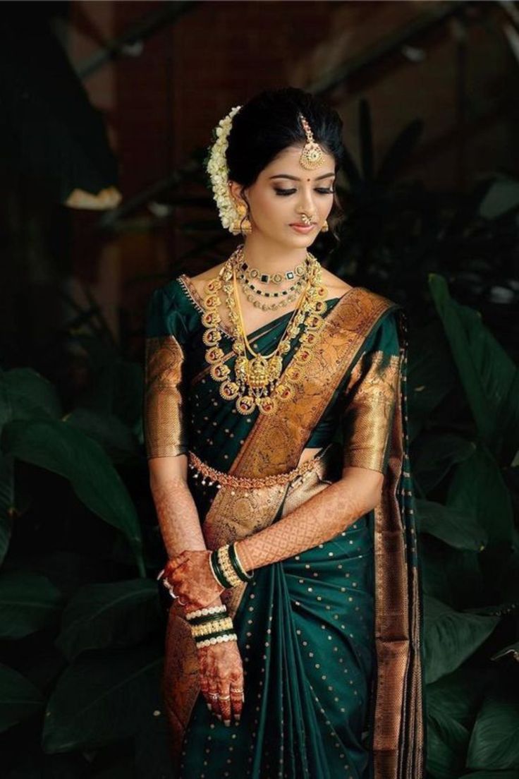 a woman in a green and gold sari with jewelry on her neck, standing next to some plants