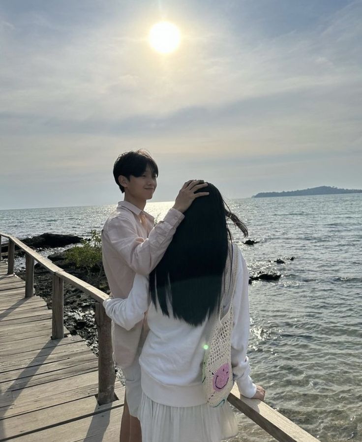 a man and woman standing on a pier next to the ocean holding each other's hair