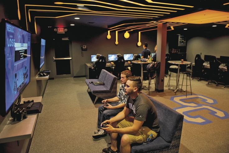 two young men sitting on couches playing video games in a large room with multiple television screens