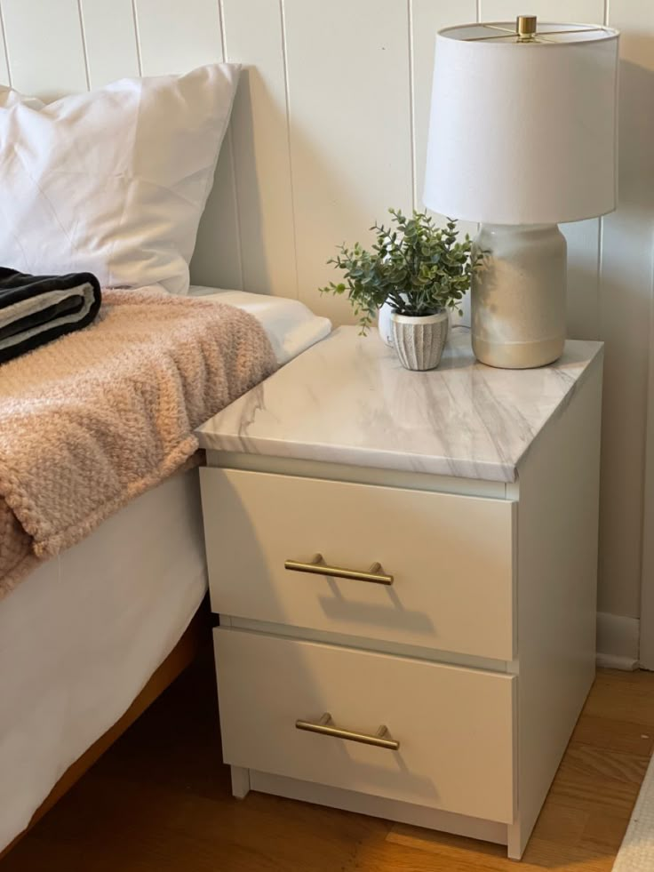 a nightstand with two drawers and a plant on it next to a bed in a bedroom