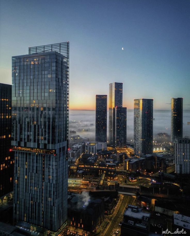 the city skyline is lit up at night with skyscrapers in the foreground and fog on the ground