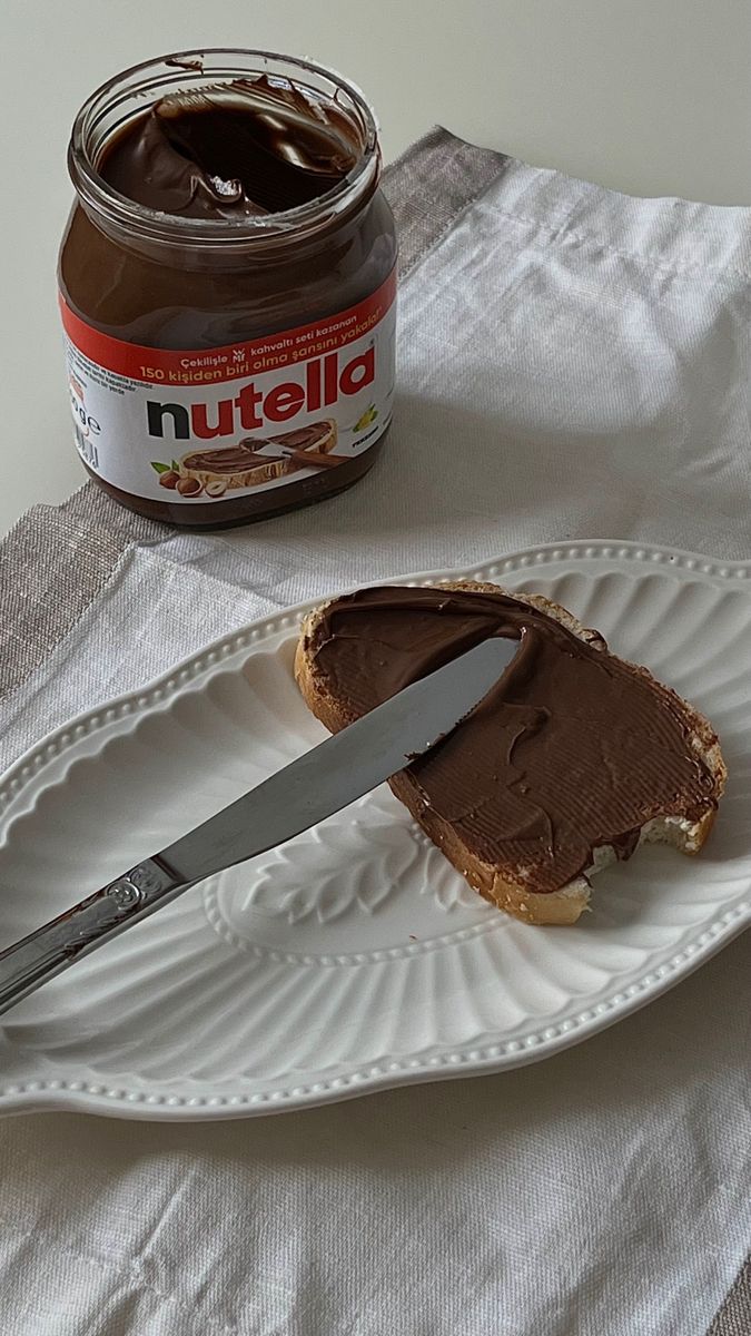 a peanut butter sandwich on a plate with a knife and jar of nutella in the background