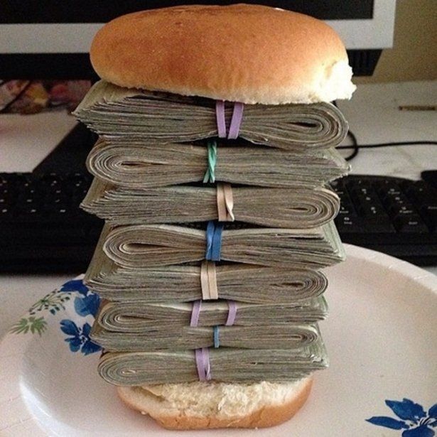 a sandwich sitting on top of a white plate covered in stacks of money next to a computer