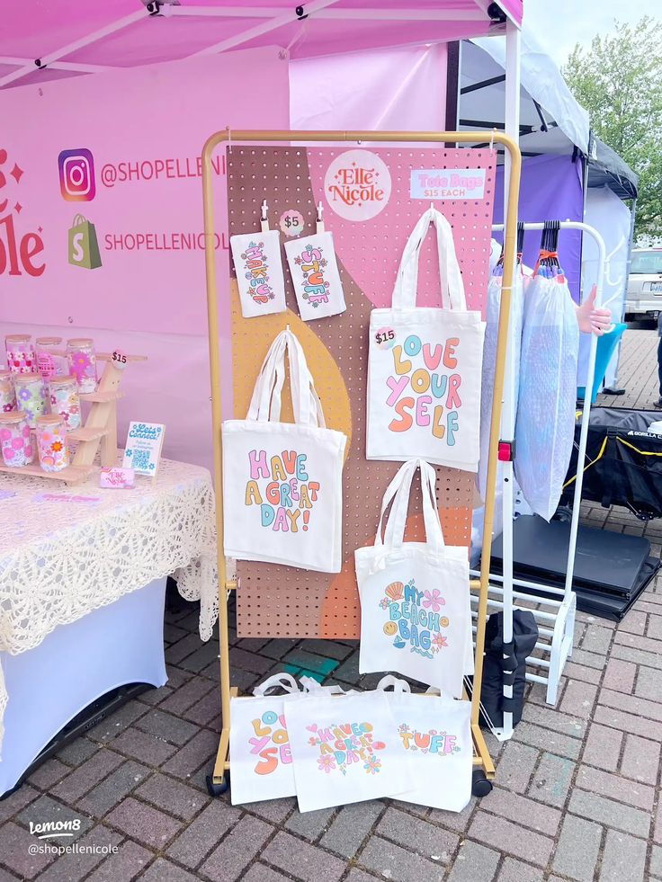 there are many bags on display under the tent at this outdoor event, including tote bags that say i love you so much