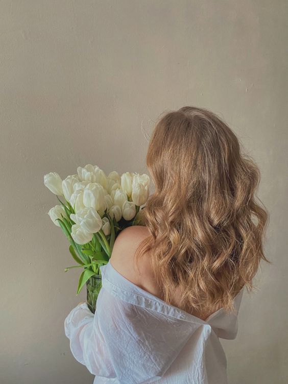 a woman holding a vase with white flowers in it's hands and looking down at the ground