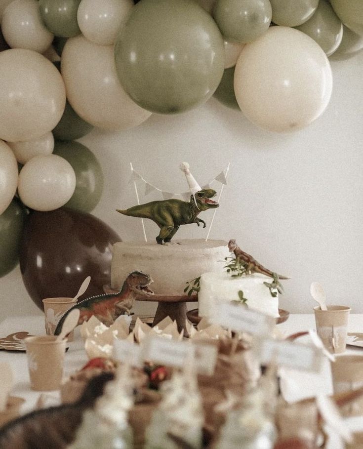 a table topped with cake and balloons in front of a dinosaur balloon wall above it