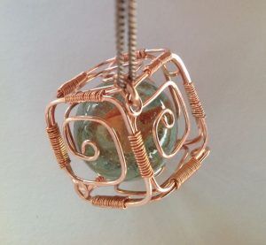 a wire wrapped pendant with a green stone in the shape of a cube hanging from a chain