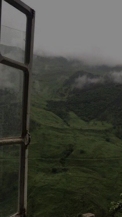 an open window in the side of a green mountain with clouds rolling over it and grass below