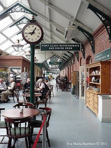 the inside of a train station with people sitting at tables, and a clock hanging from the ceiling