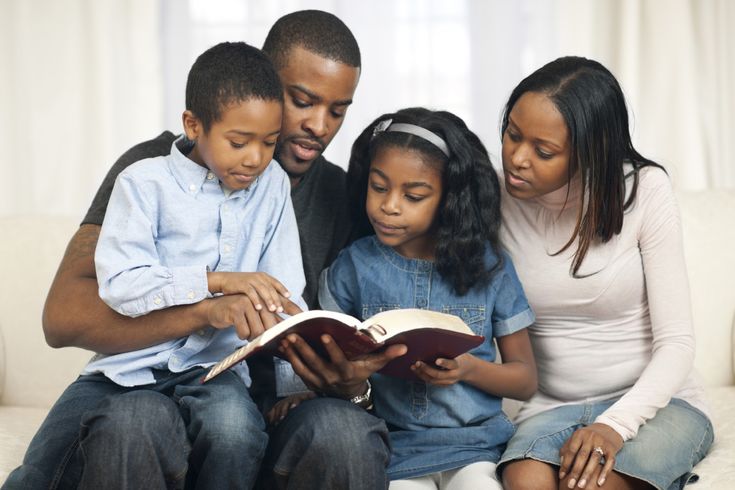 three adults and two children sitting on a couch looking at an open book while the child is reading it