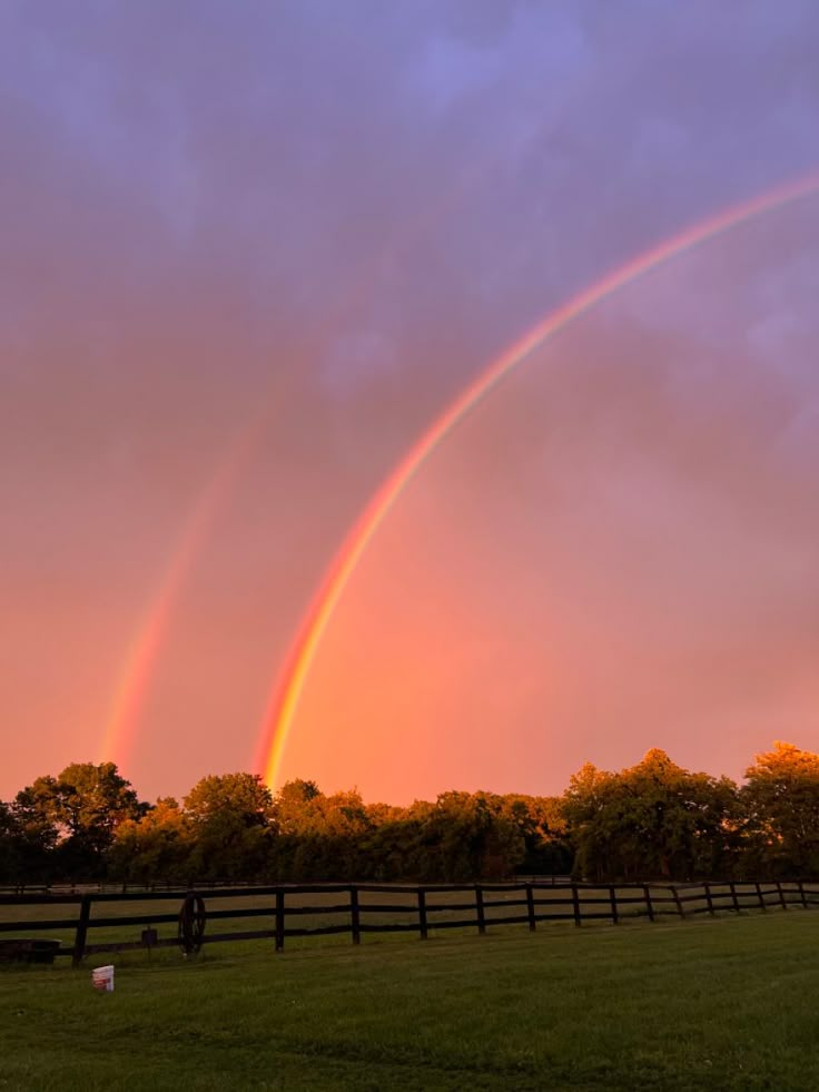 two rainbows in the sky over a grassy field with trees and a wooden fence