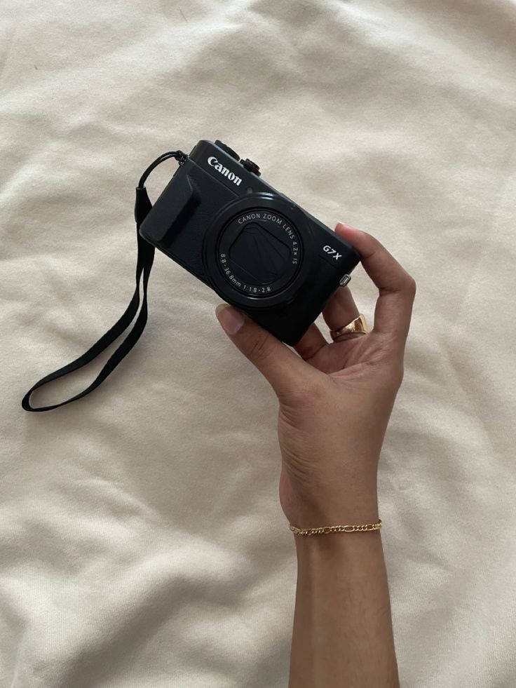 a hand holding a black camera on top of a white bed sheet with a gold chain around it