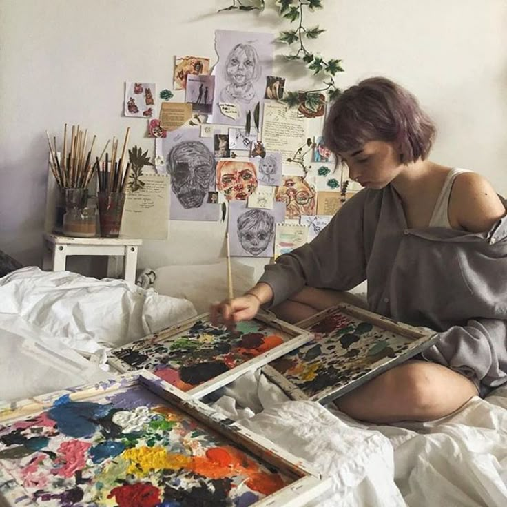 a woman sitting on top of a bed next to an easel filled with paintings