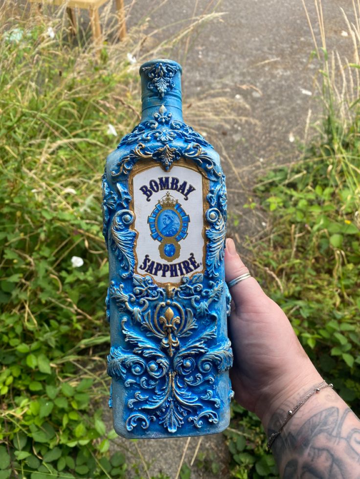 a hand holding a blue bottle with the words bobby's sapphire on it in front of some bushes