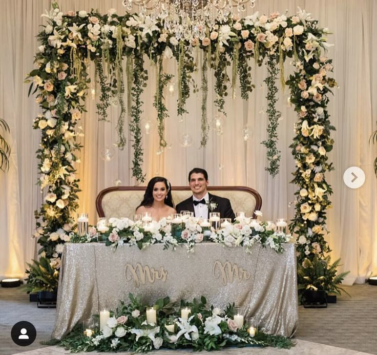 two people sitting at a table with flowers and greenery