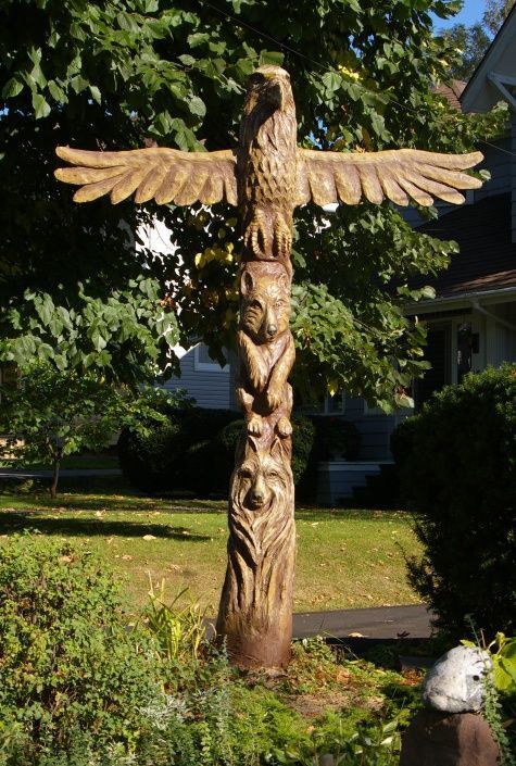 a carved wooden bird on top of a pole in front of a tree and bushes