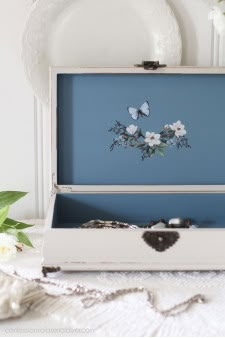 an open white box sitting on top of a table next to some flowers and butterflies