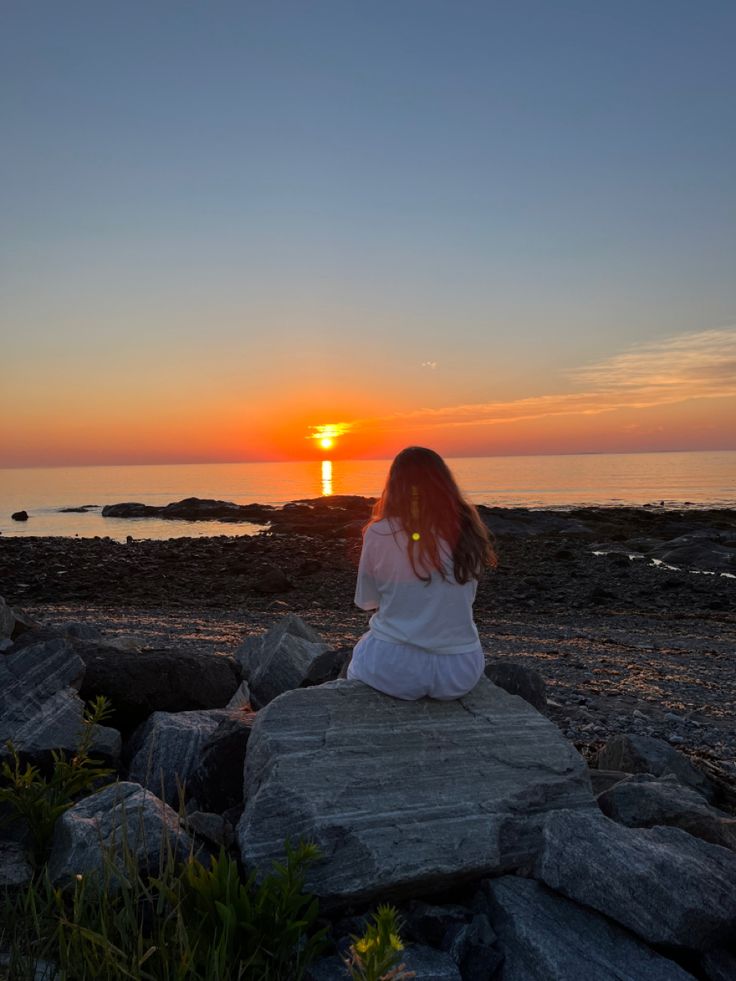 a woman sitting on top of a rock near the ocean at sunset with her back to the camera