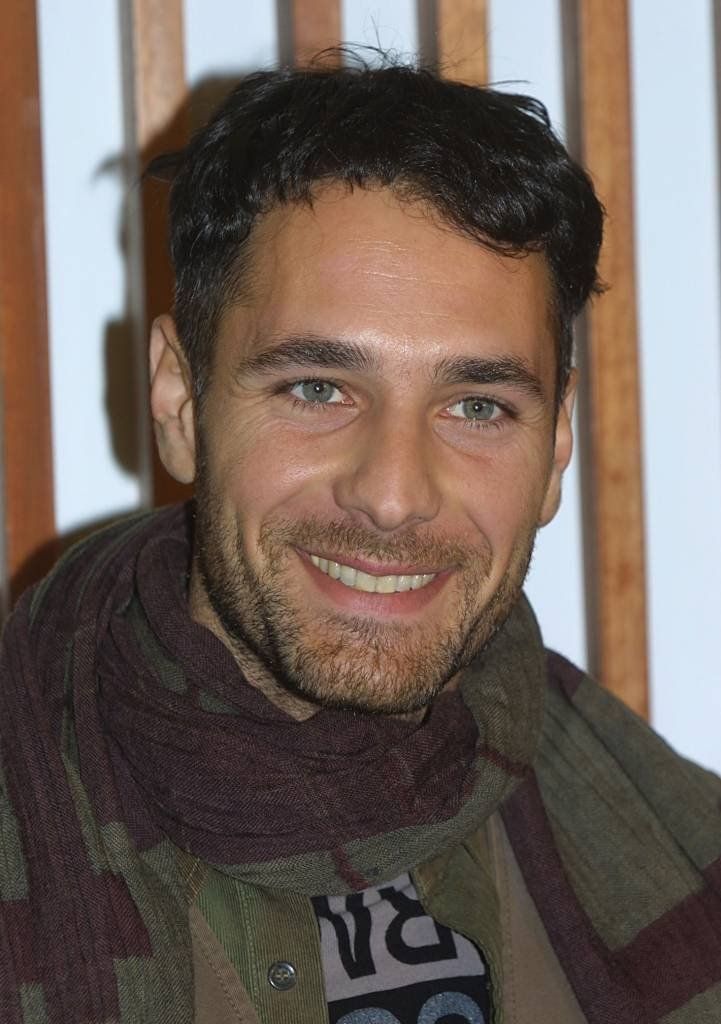 a smiling man wearing a scarf in front of a wooden wall with vertical slats