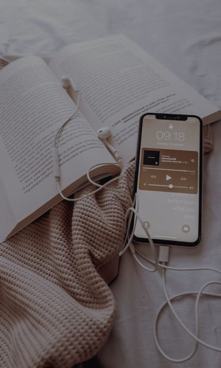 an open book and headphones are laying on a bed next to an iphone that is plugged in