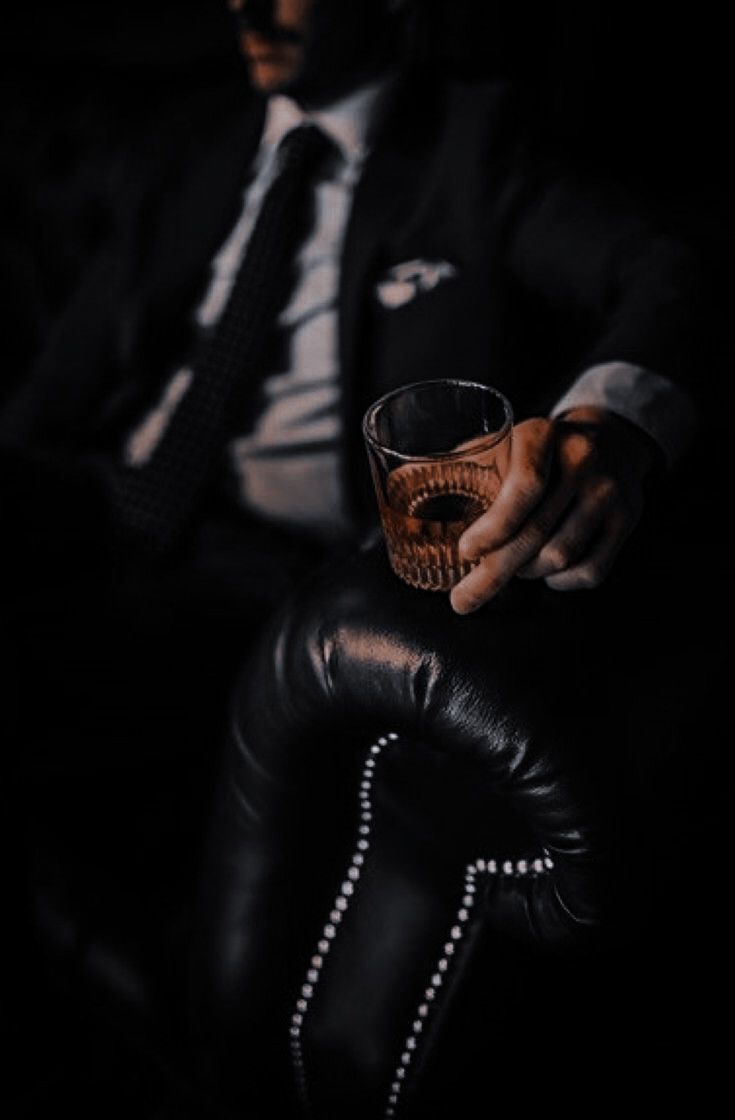 a man sitting in a chair holding a glass filled with liquid and wearing a suit