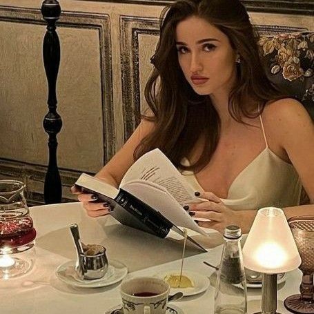 a woman sitting at a table with a book in her hand and wine glasses on the table