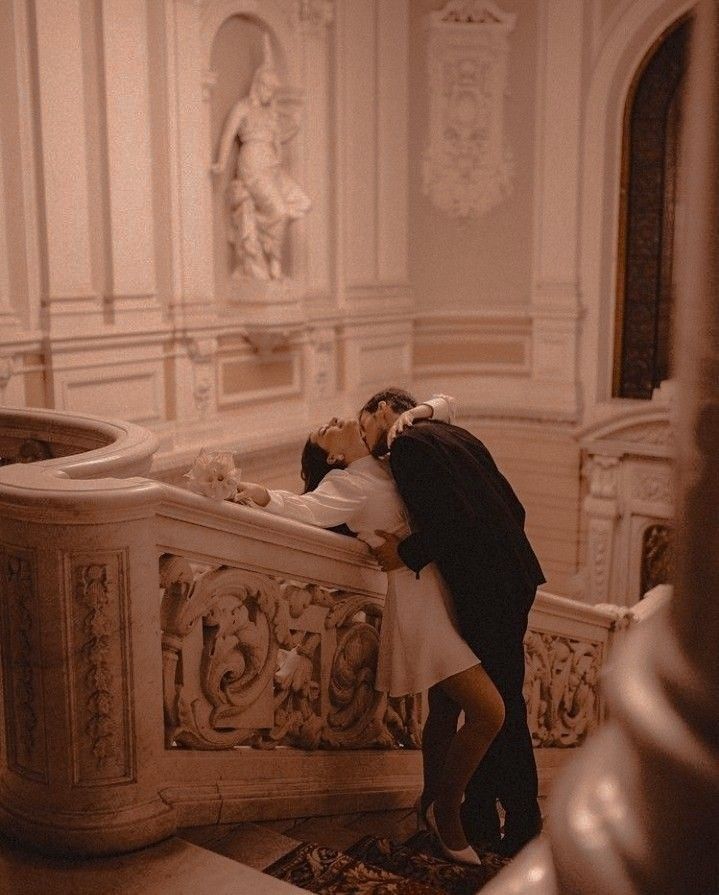 a man and woman are kissing on the stairs in an ornate room with white walls