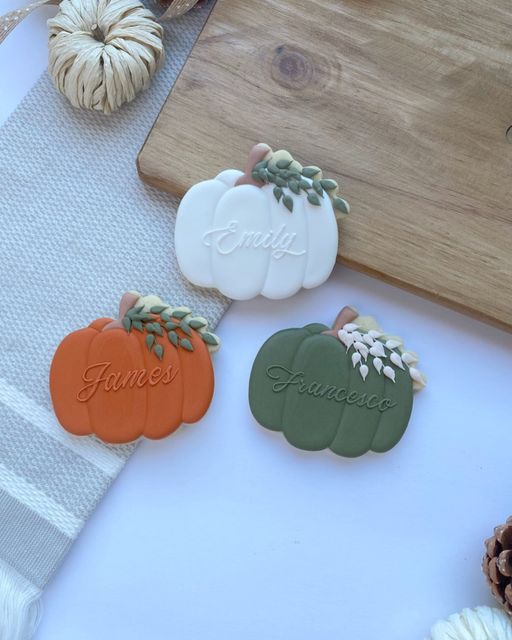 three decorated cookies sitting on top of a wooden cutting board next to pine cones and pumpkins