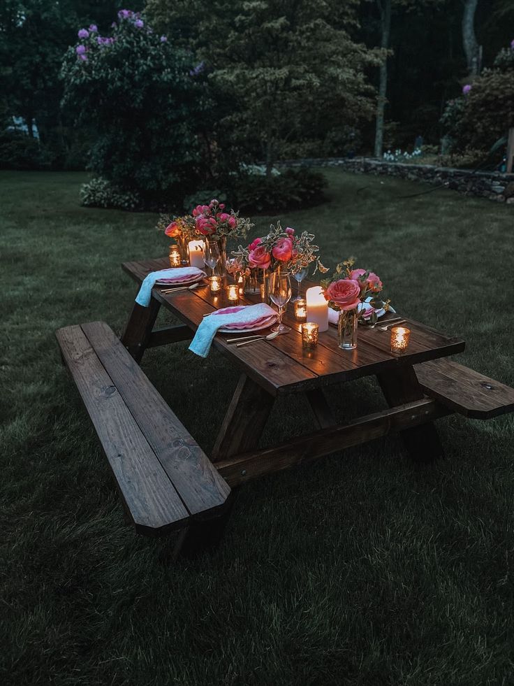 a picnic table with candles and flowers on it in the grass at night, surrounded by greenery