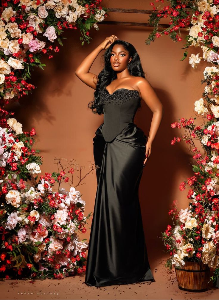 a woman in a long black dress posing for a photo with flowers on the wall behind her