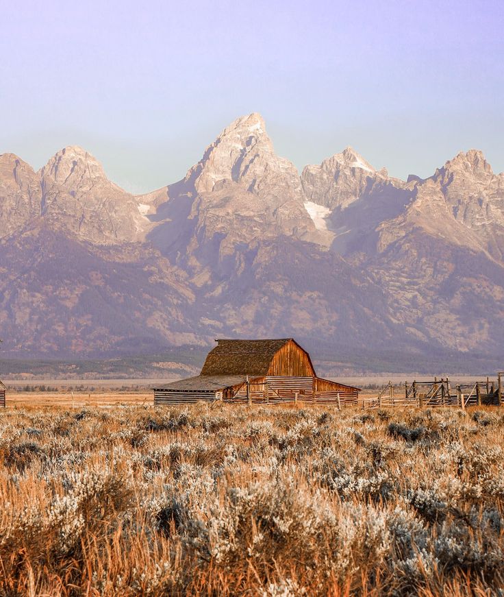 an old barn in the middle of a field with mountains in the background