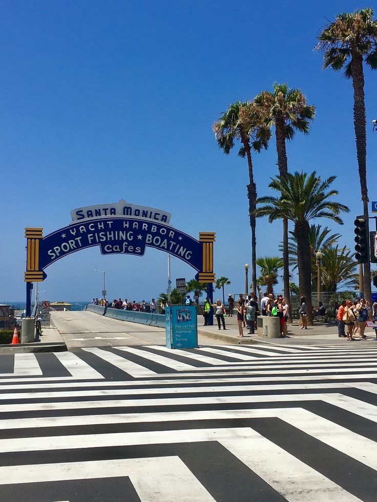 people are walking on the beach near some palm trees and a blue sign that says santa monica