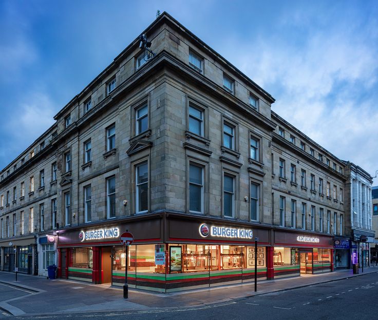 an old building on the corner of a city street at dusk with shops lit up