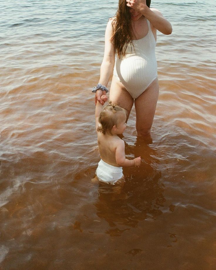 a woman in a white bodysuit is standing in the water with a baby on her hip