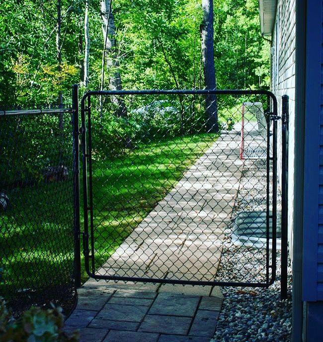 an open gate in front of a house on a path between two buildings with trees behind it