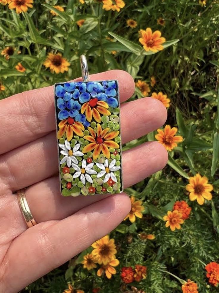 a hand holding a small square pendant with flowers on it's side and yellow daisies in the background