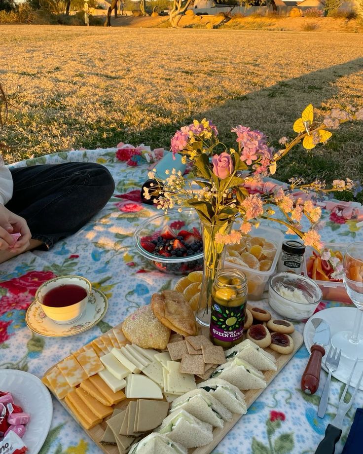 a woman sitting at a picnic table with food and drinks on it, in the middle of a field