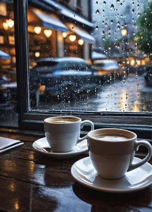 two cups of coffee sit on a table in front of a window with raindrops