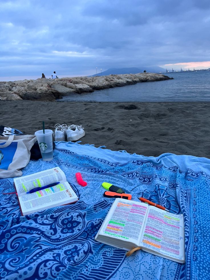 an open book sitting on top of a blue blanket next to the ocean with writing utensils