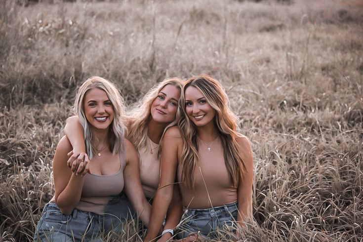 three beautiful young women sitting next to each other in a grassy field with tall grass