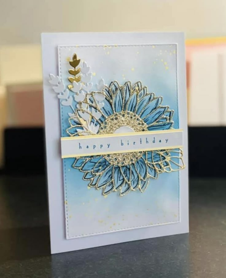 a blue and white birthday card with gold foil flowers on it, sitting on a table