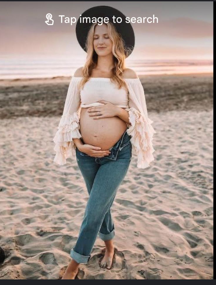 a pregnant woman wearing jeans and a hat on the beach with her belly exposed, standing in front of the ocean