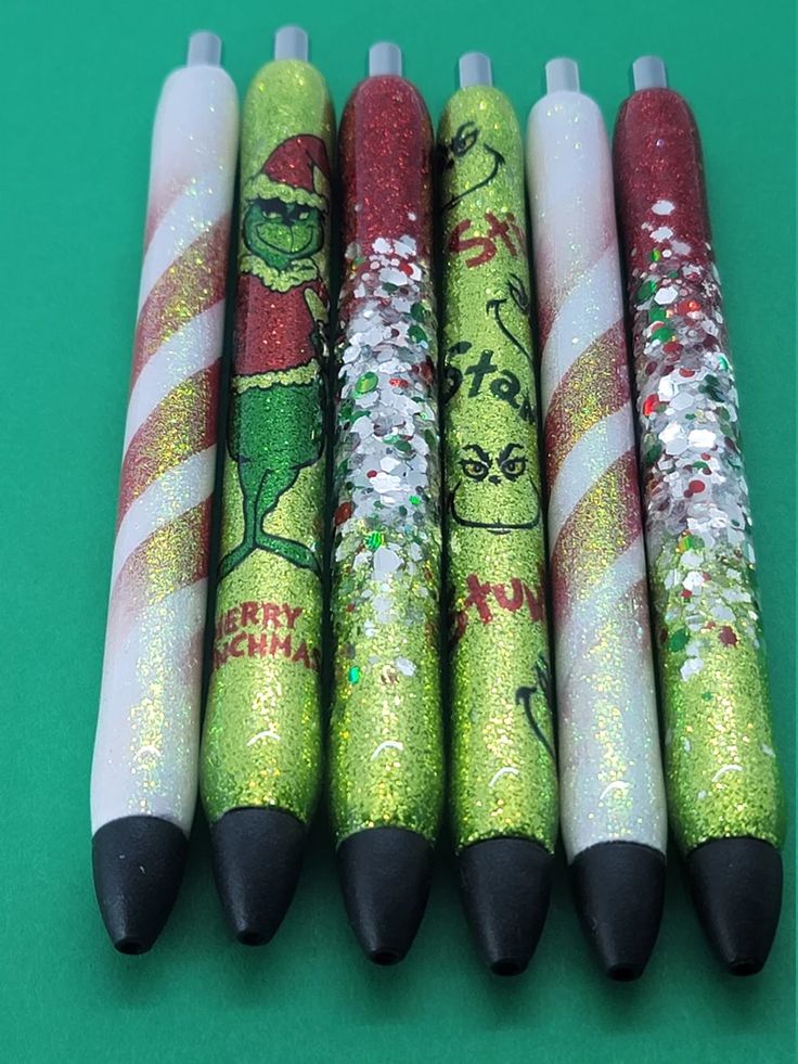 four christmas themed pens are lined up on a green surface with white and red glitters