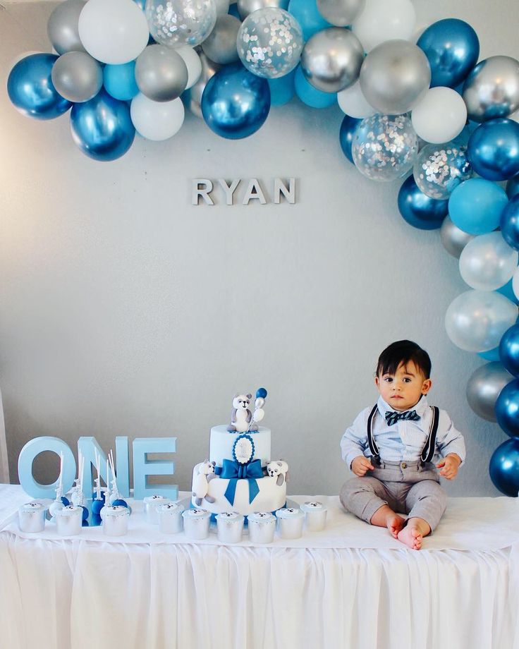 a baby boy sitting on top of a table next to a blue and white cake