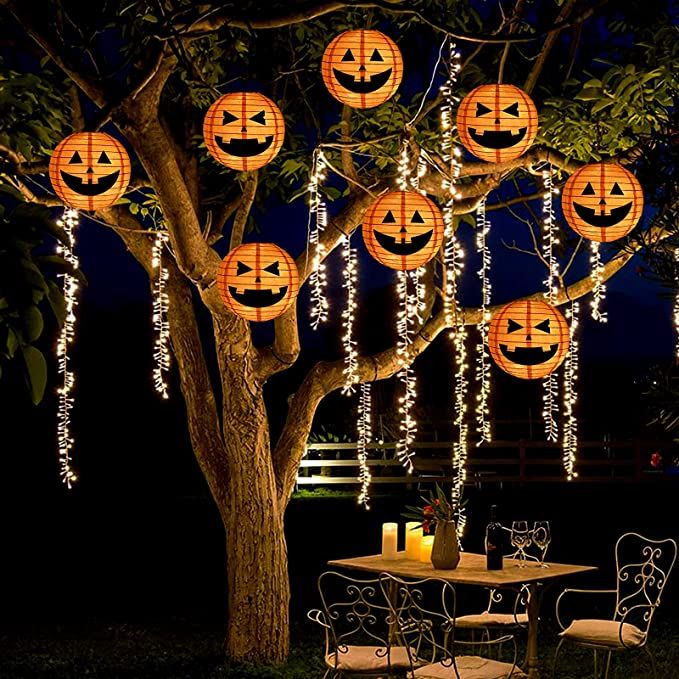 an outdoor table and chairs under a tree decorated with lanterns for the halloween season at night