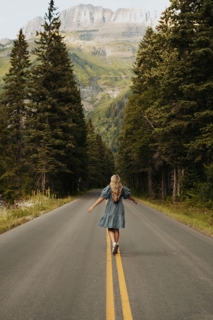 a woman is walking down the middle of an empty road in front of some trees