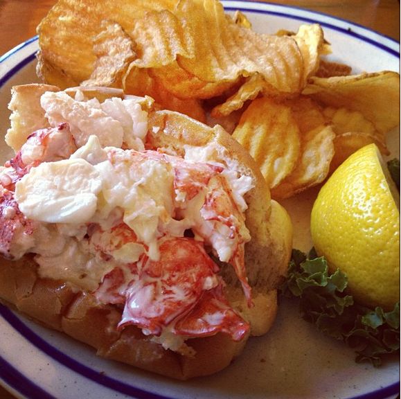 a lobster sandwich and potato chips on a plate with lemon wedges next to it